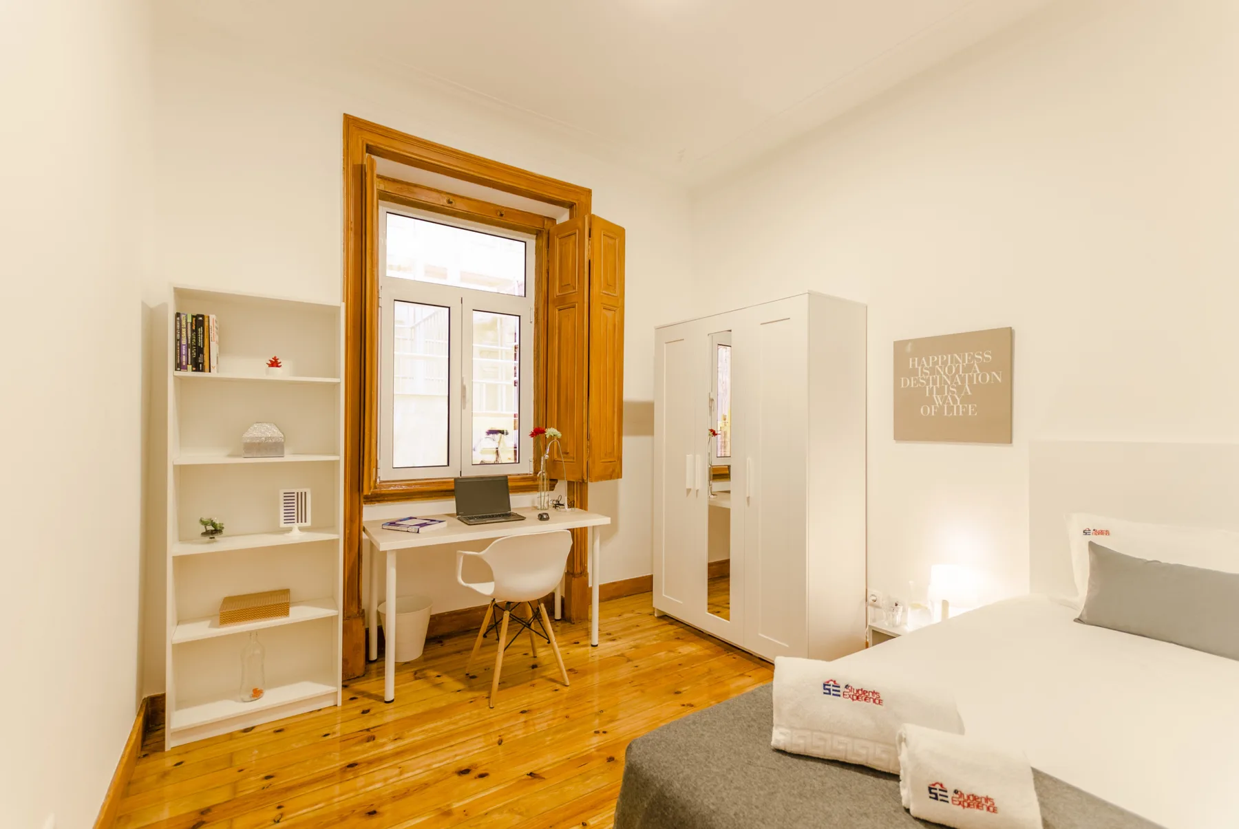 Un dormitorio luminoso con un escritorio junto a una ventana alta de madera y una cama.