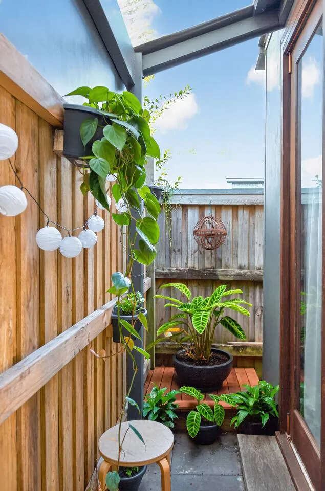 A narrow outdoor patio has potted greenery, wooden fencing, and a small stool.