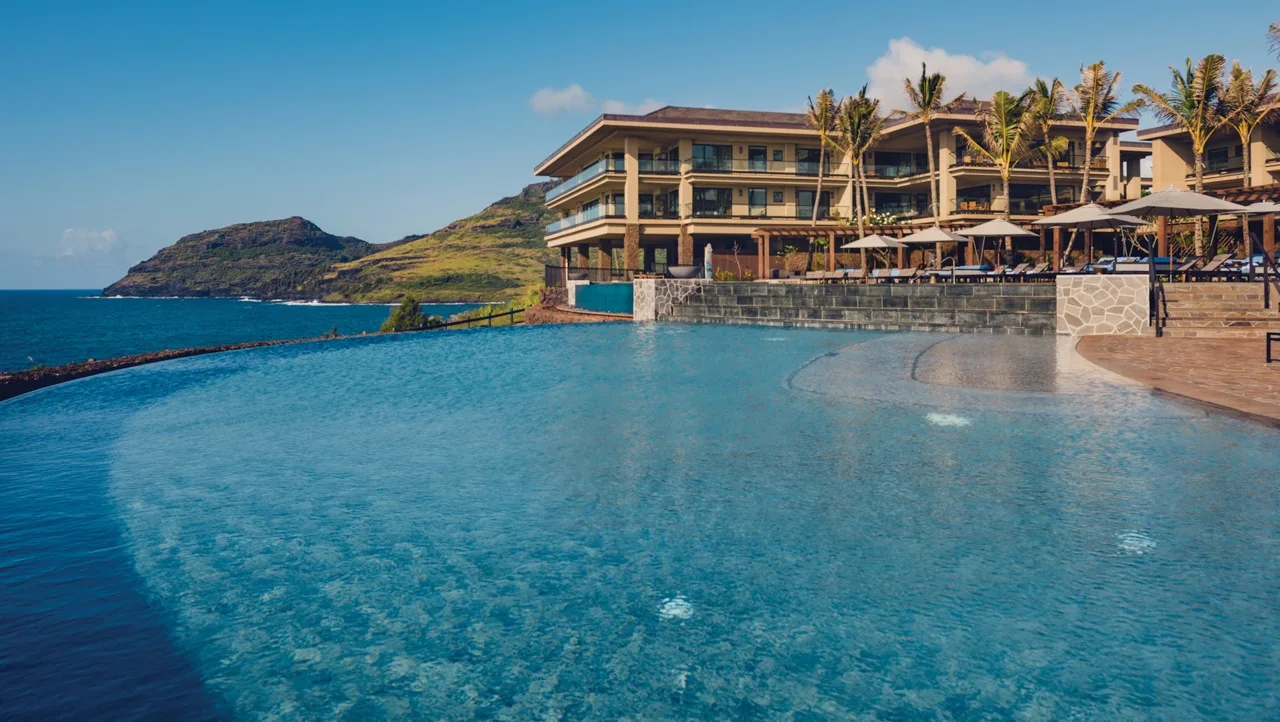 An outdoor swimming pool overlooks the ocean beside a palm-lined resort building.