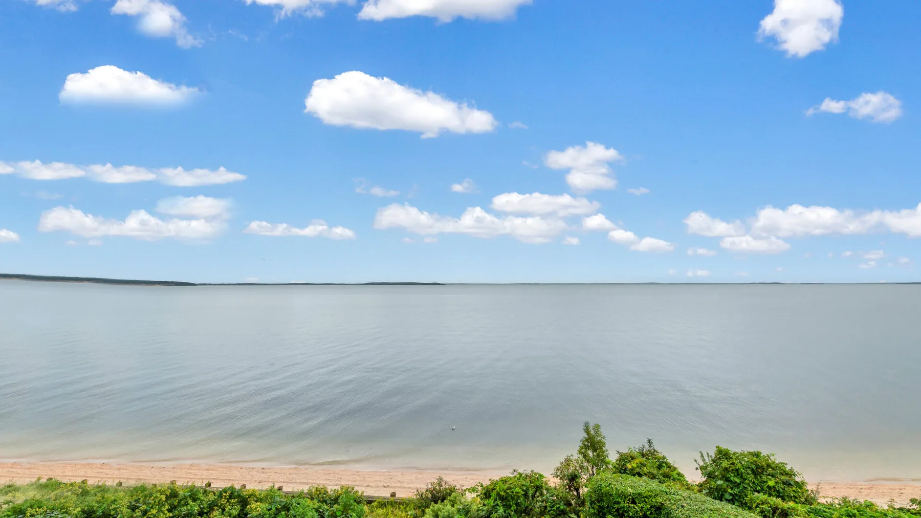 Una amplia vista junto al agua muestra aguas tranquilas bajo un cielo azul con nubes.