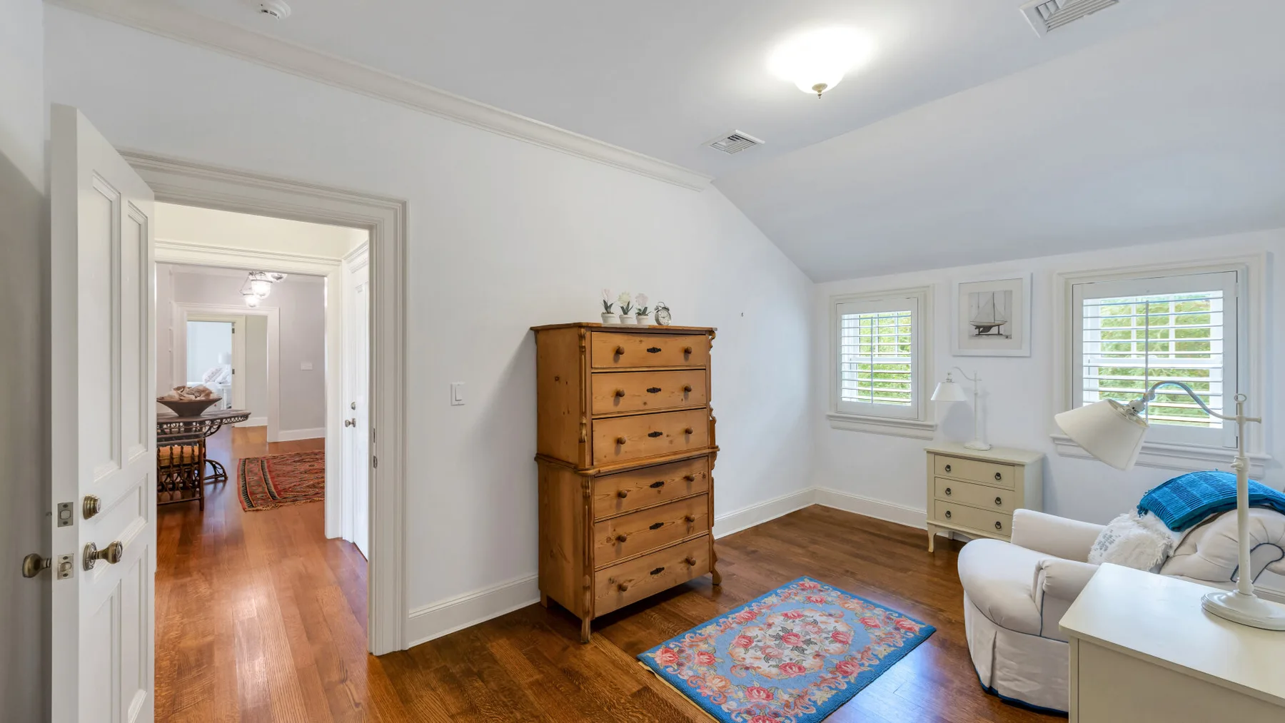 Un dormitorio luminoso con una cómoda de madera, dos ventanas y una alfombra estampada.