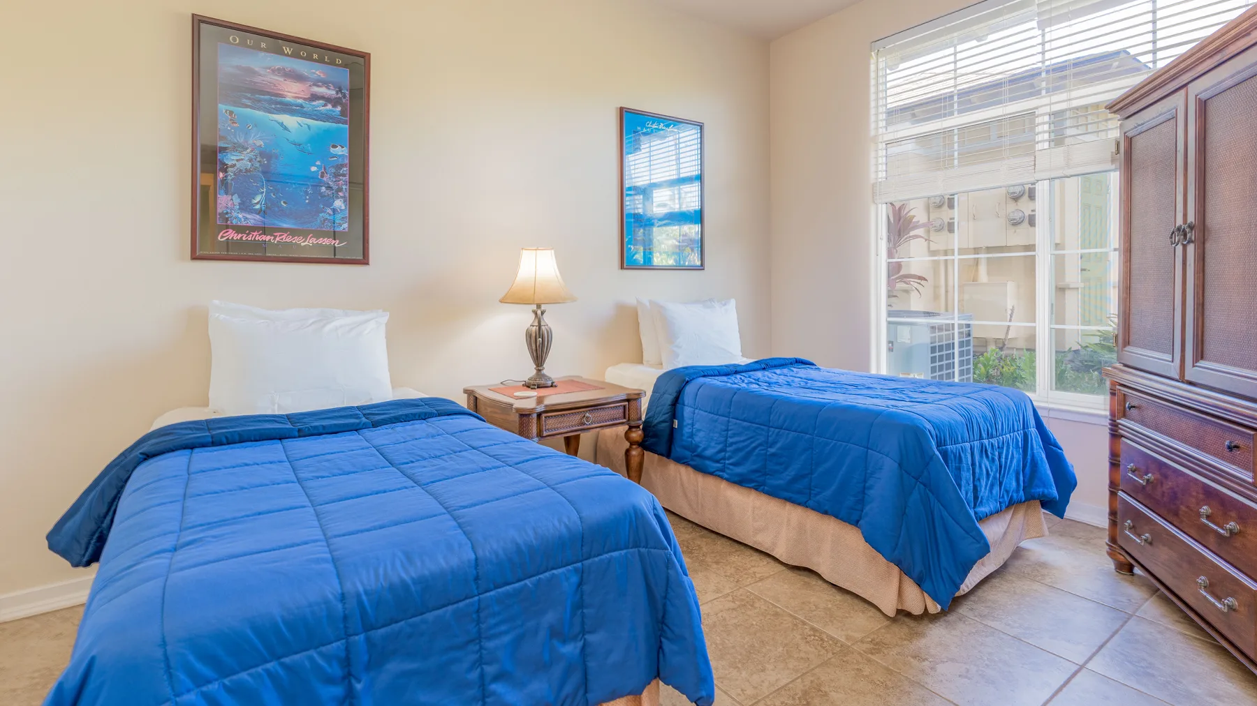 A bedroom with two beds covered in blue bedding beside a bright window.