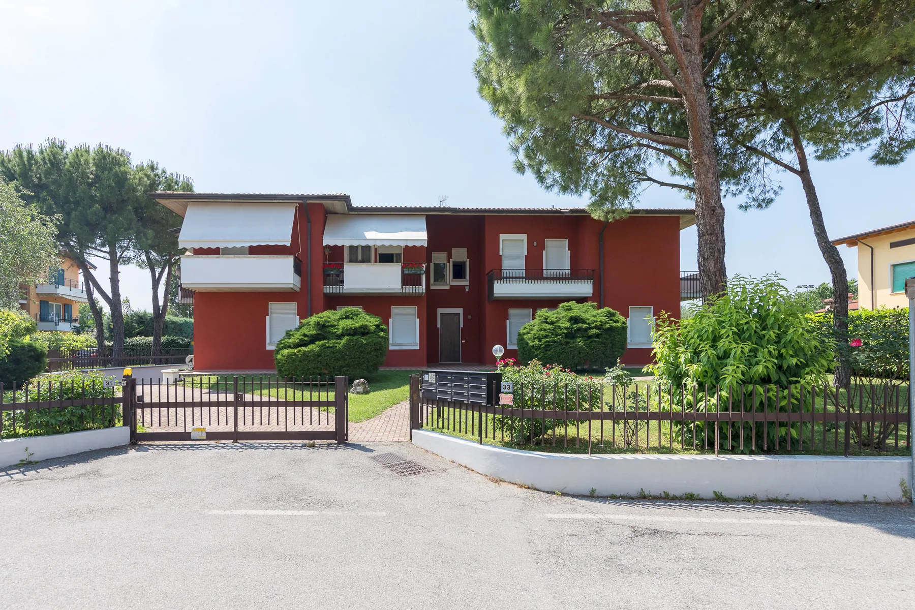 A red two-story building stands behind a gated entrance, surrounded by trees and shrubs.