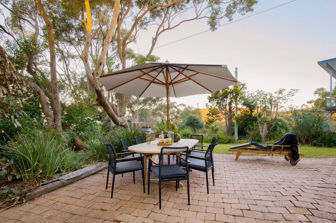An outdoor dining area with a large umbrella, surrounded by trees and garden plants.