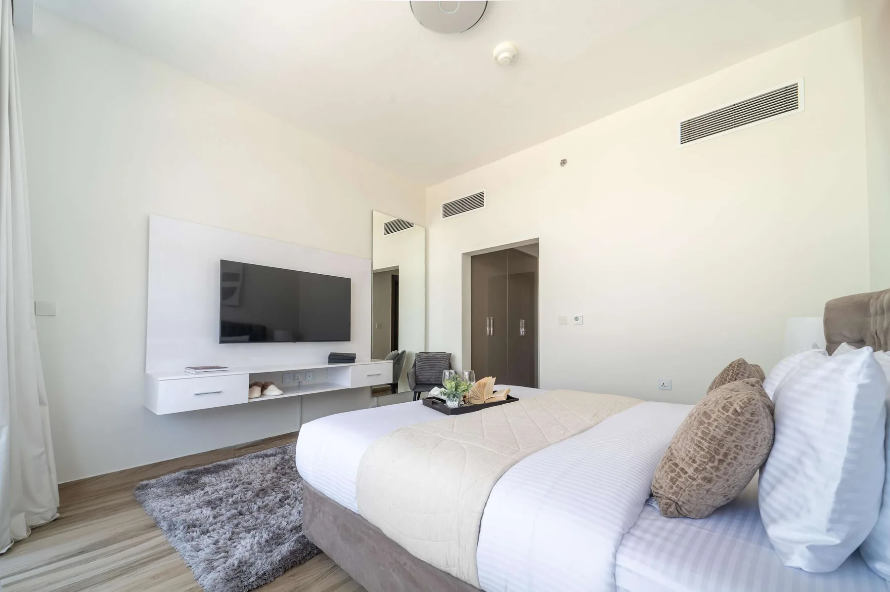 Un dormitorio luminoso con una cama, un televisor montado en la pared y suelo de madera clara.