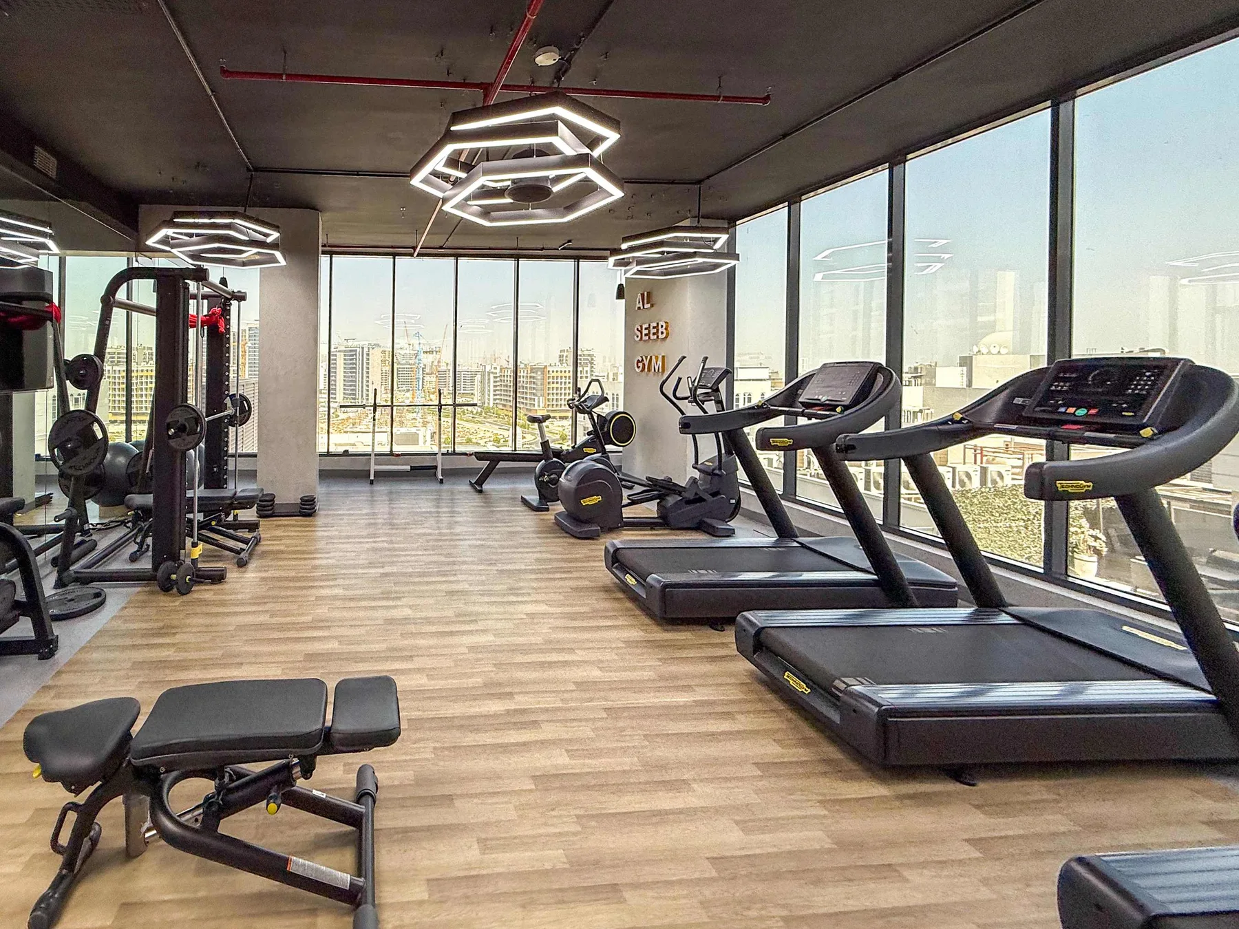 Un gimnasio con equipos de ejercicio y grandes ventanales con vistas a la ciudad.