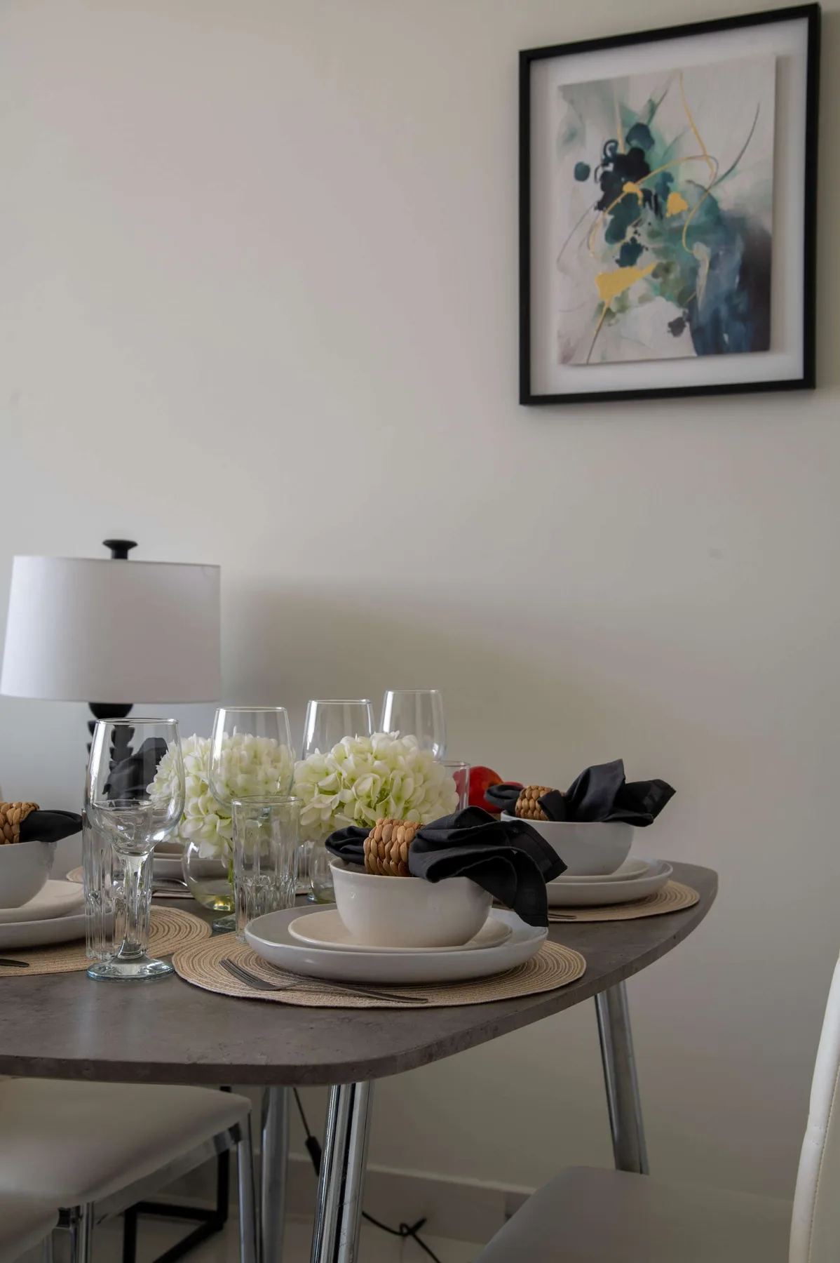 Una mesa de comedor está puesta con vajilla, cristalería, flores y una lámpara bajo un cuadro enmarcado.