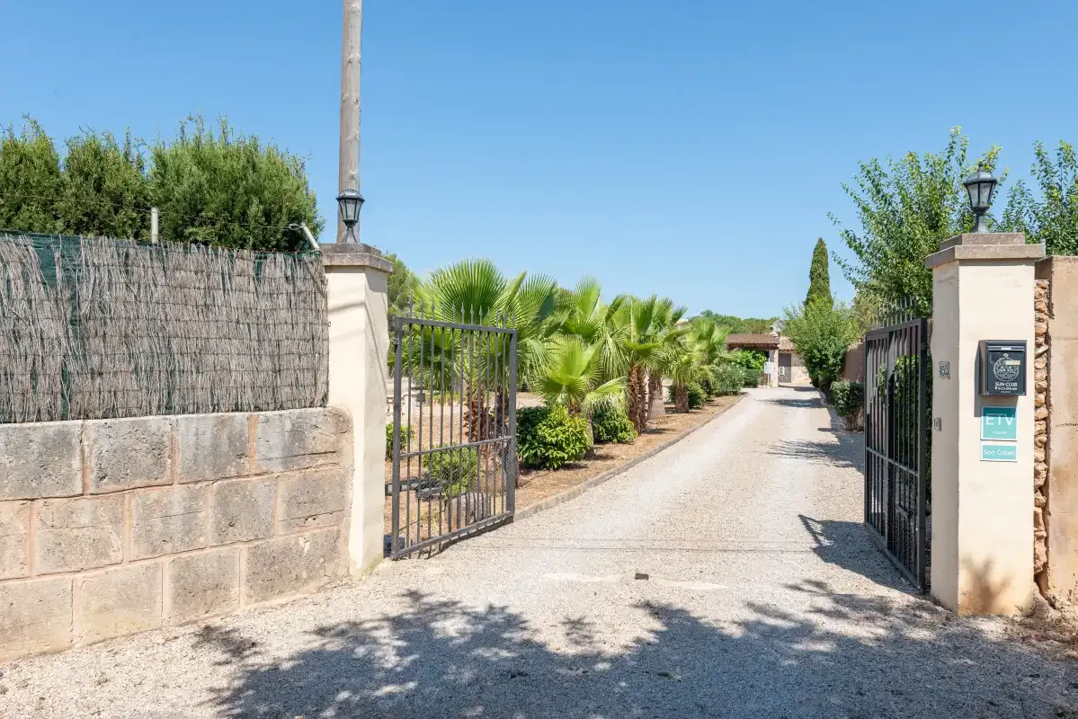 A gravel driveway leads through an open gate toward palm trees and buildings.