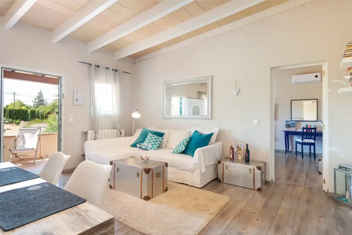 A bright living room has white seating, a low table, and an open doorway to a dining area.