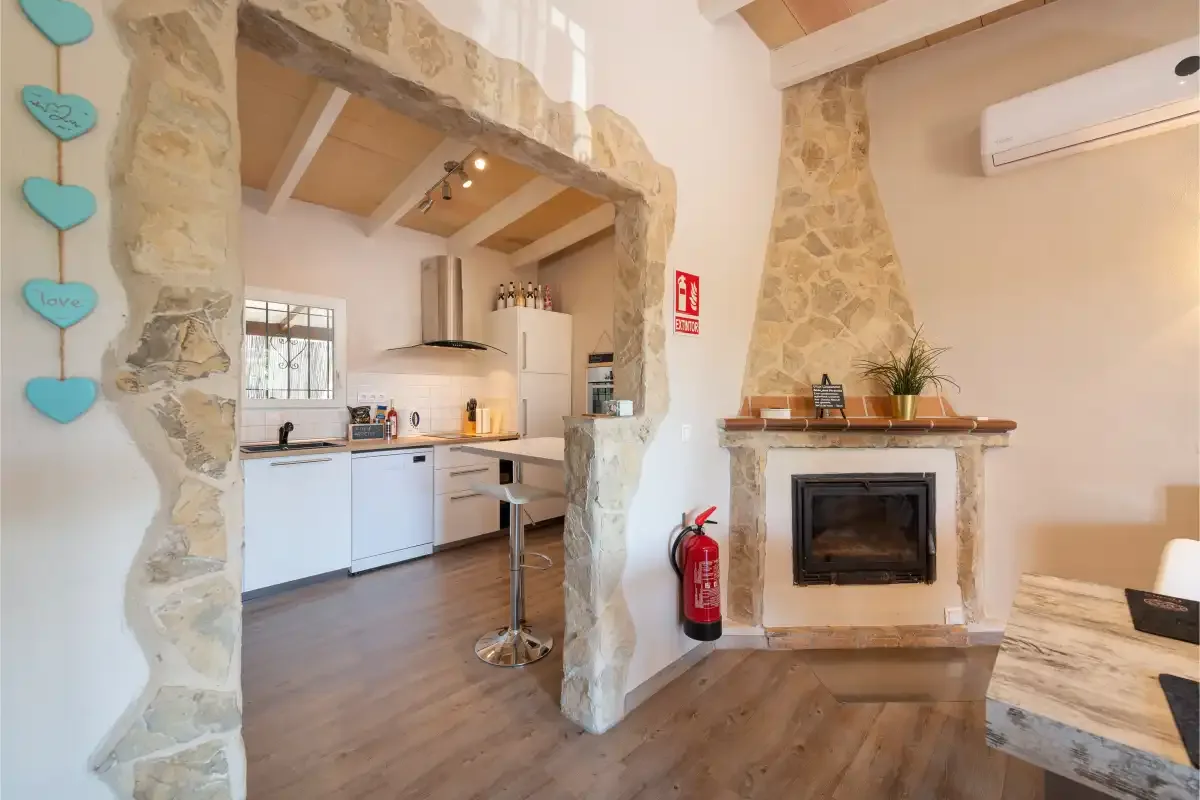 A rustic living area opens onto a kitchen, with a stone fireplace beside it.