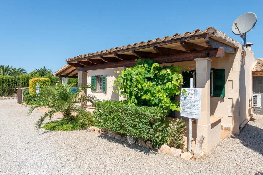 A single-story house with a tiled roof, shaded porch, and leafy plants.