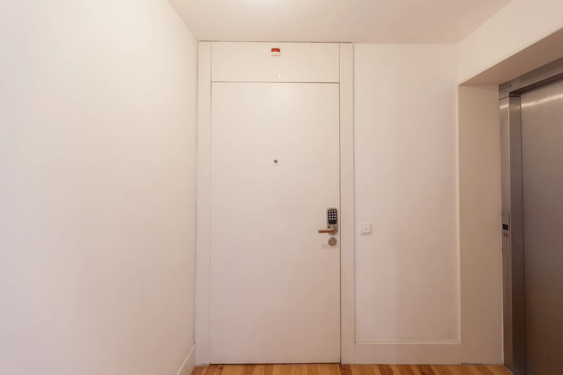 A white apartment entrance door opens onto a hallway with wood flooring.