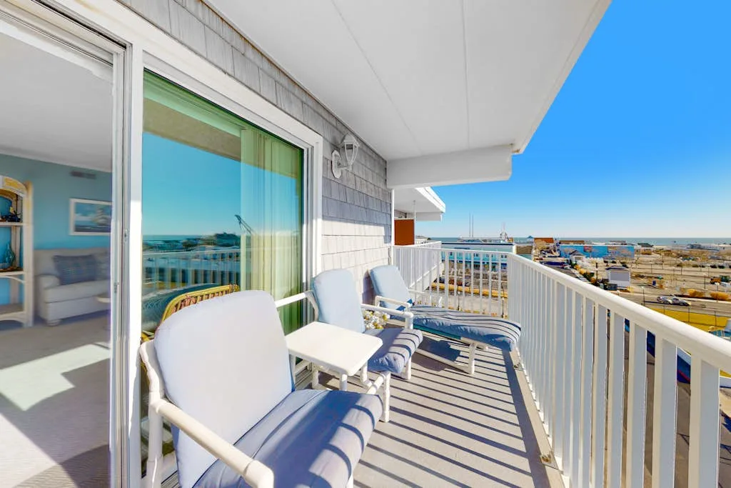 A balcony with outdoor seating overlooks the ocean and nearby buildings.