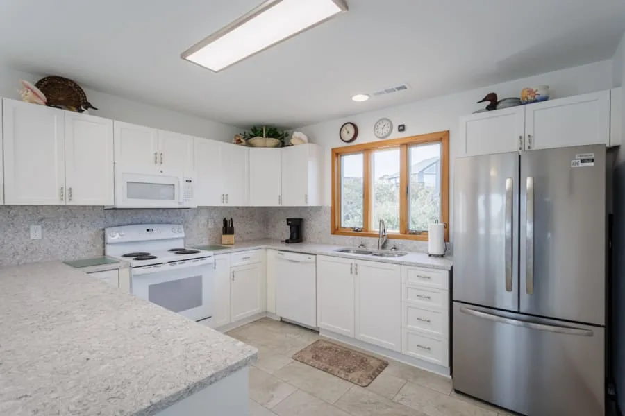 Una cocina blanca y luminosa con un fregadero bajo una ventana con marco de madera.
