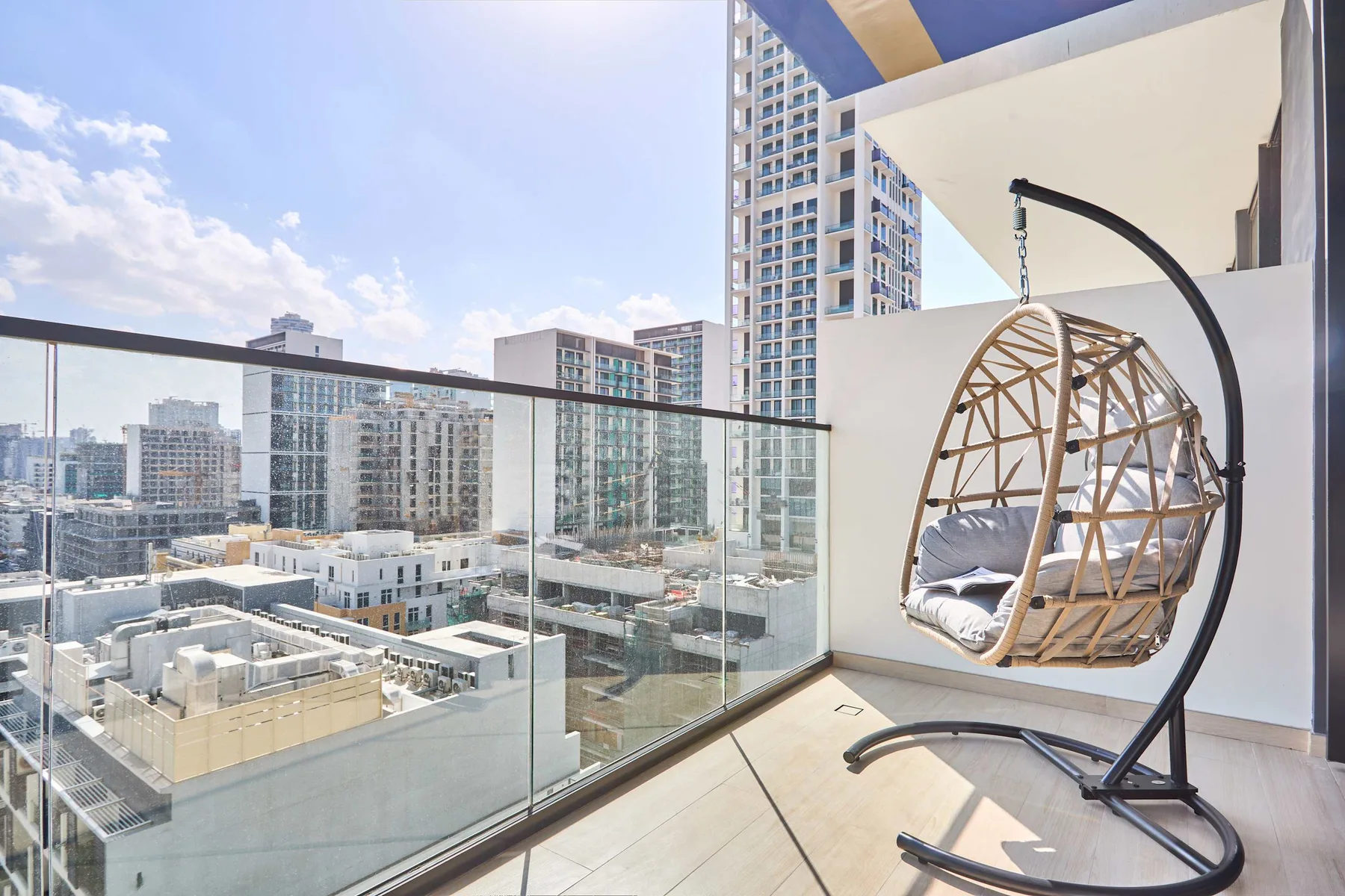 A balcony with glass railing and a hanging chair overlooks nearby city buildings.
