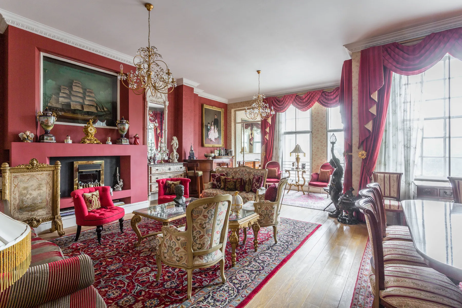 A formal living room with red walls, patterned rugs, and tall curtained windows.