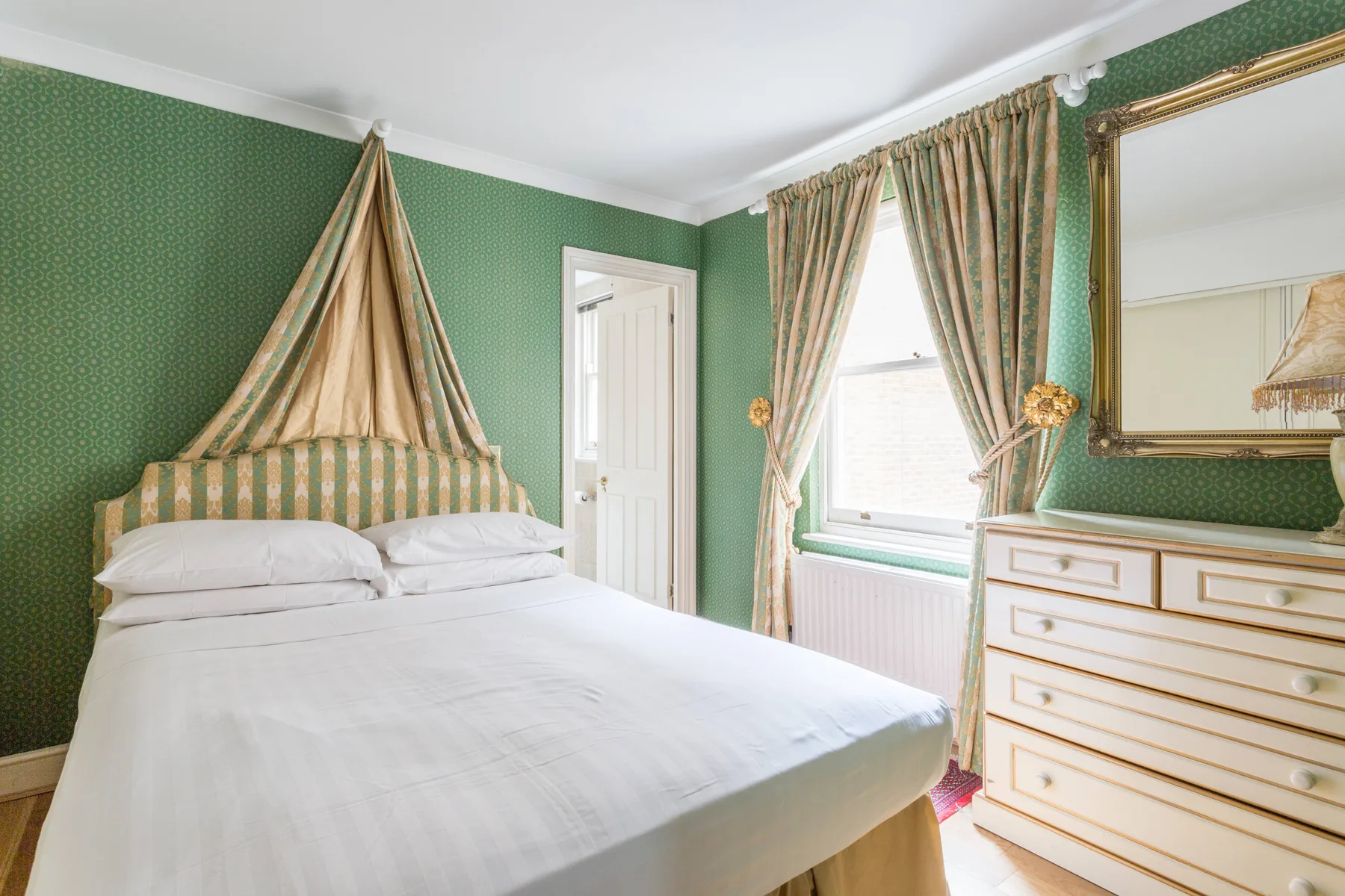 Un dormitorio con una cama a rayas, paredes verdes estampadas y una cómoda bajo un espejo.