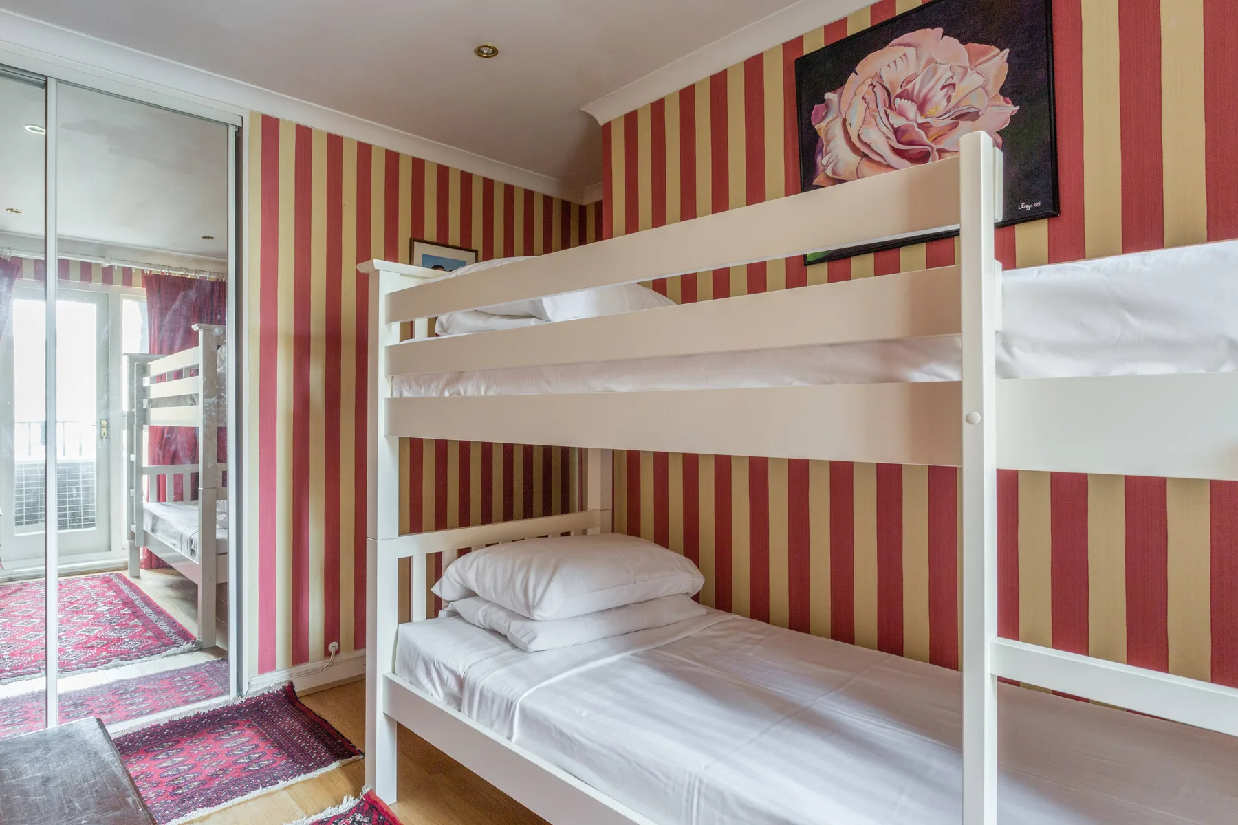 Un dormitorio con literas blancas, papel pintado a rayas y un cuadro floral.
