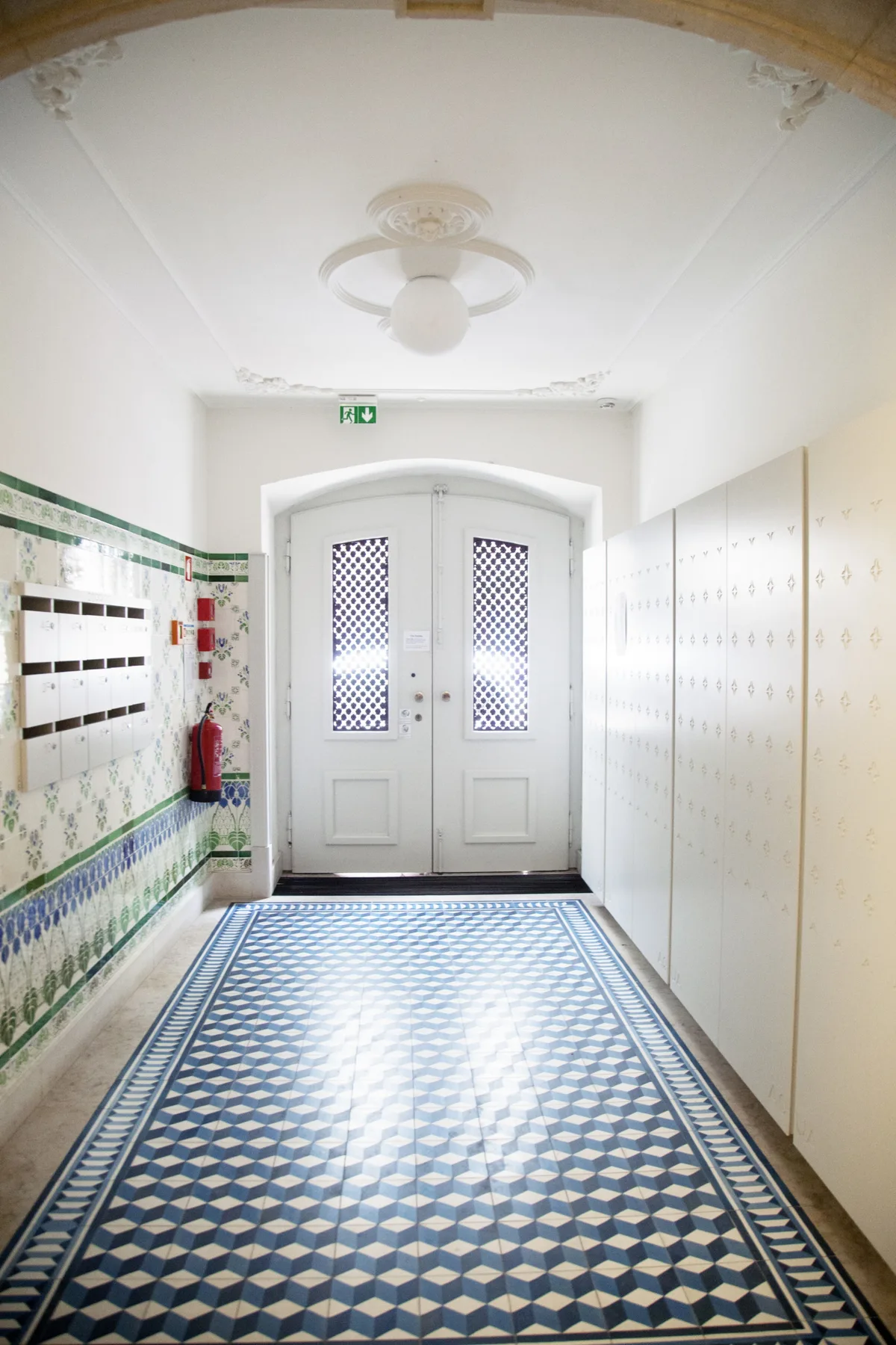 A tiled apartment hallway leads to a pair of white doors.