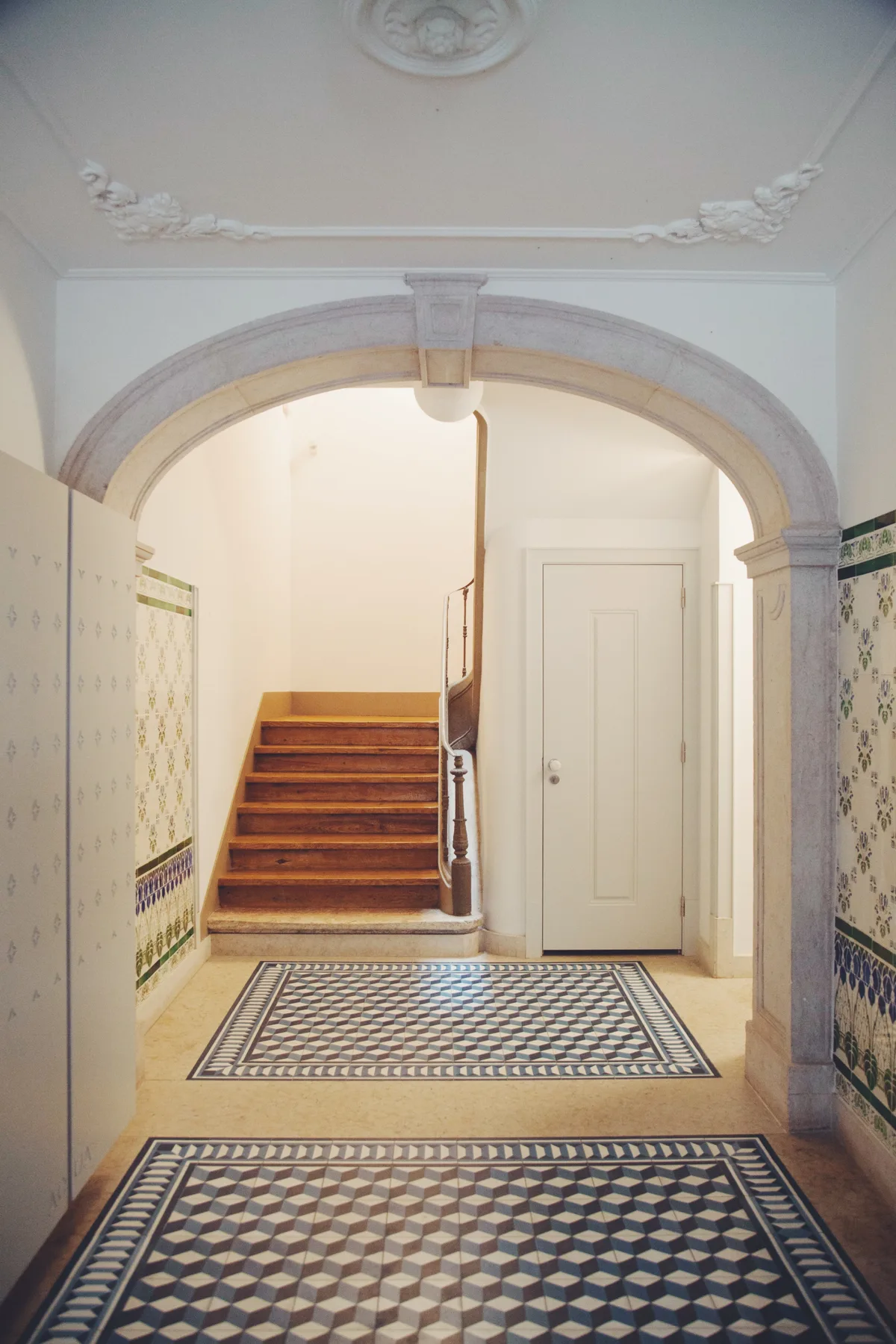 An ornate tiled hallway leads through an arch to a staircase and closed door.