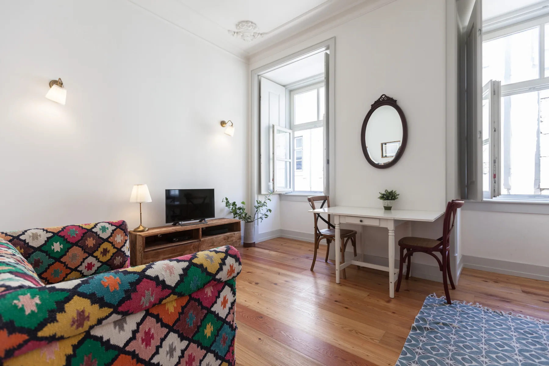 A living room with colorful patterned seating and a small dining area by the windows.
