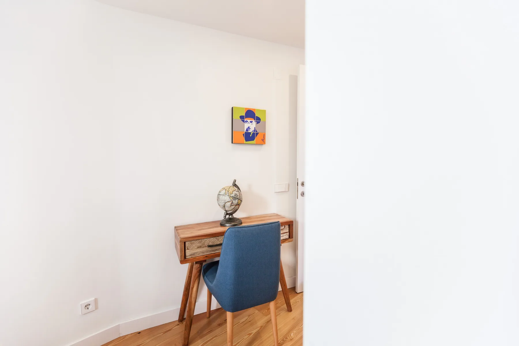 Un escritorio compacto y un asiento azul están junto a una pared blanca en una habitación luminosa.