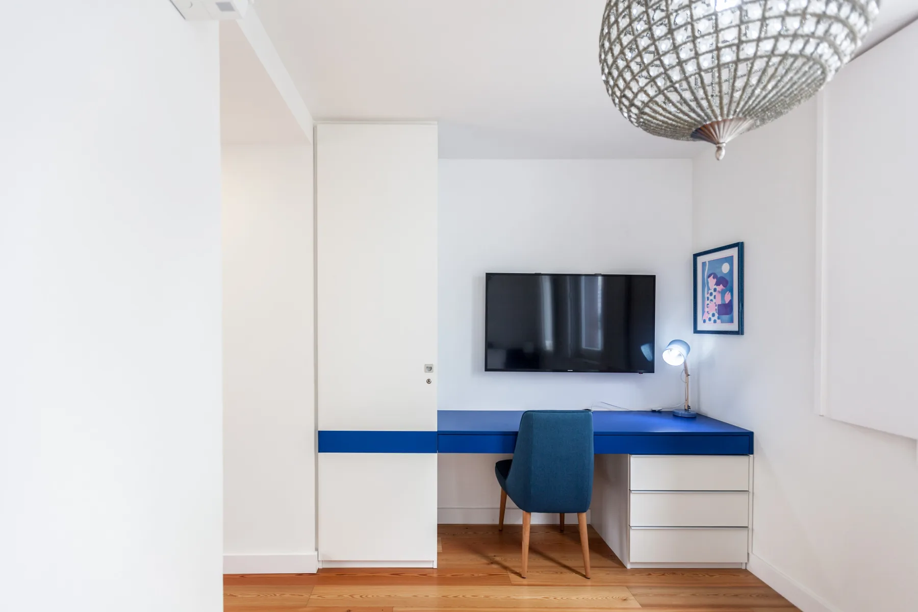 Una habitación luminosa con escritorio, silla, televisor en la pared y una obra enmarcada.