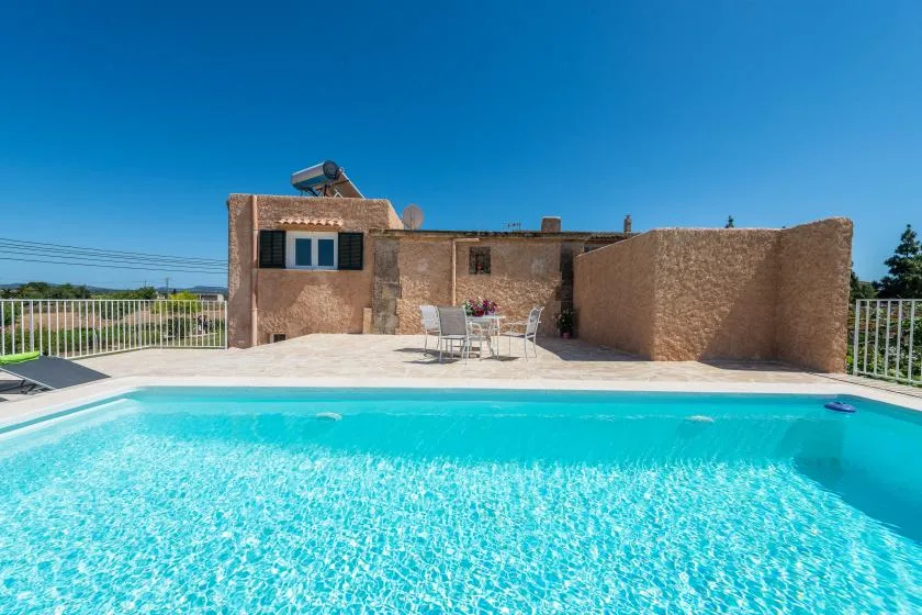 An outdoor swimming pool sits beside a sunlit stone building and patio seating.