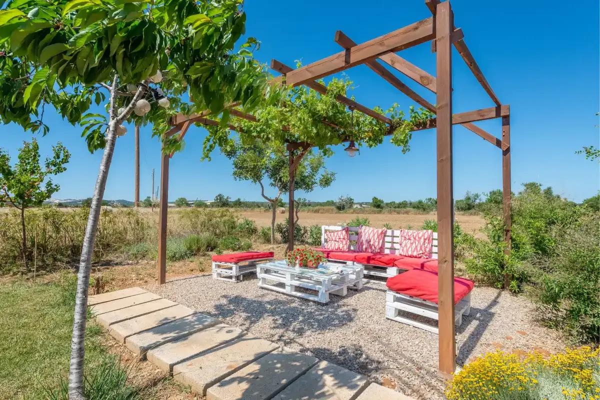 An outdoor seating area beneath a wooden pergola, with red cushions and a low table.