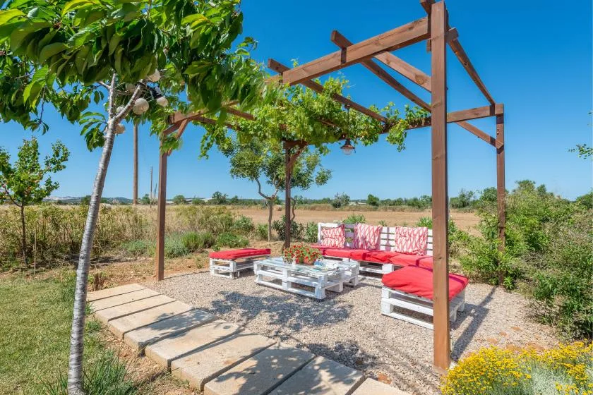 An outdoor seating area beneath a wooden pergola, with red cushions and low tables.
