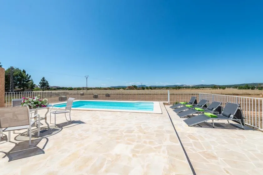 A sunny pool terrace has lounge seating, a dining table, and open views beyond the railing.
