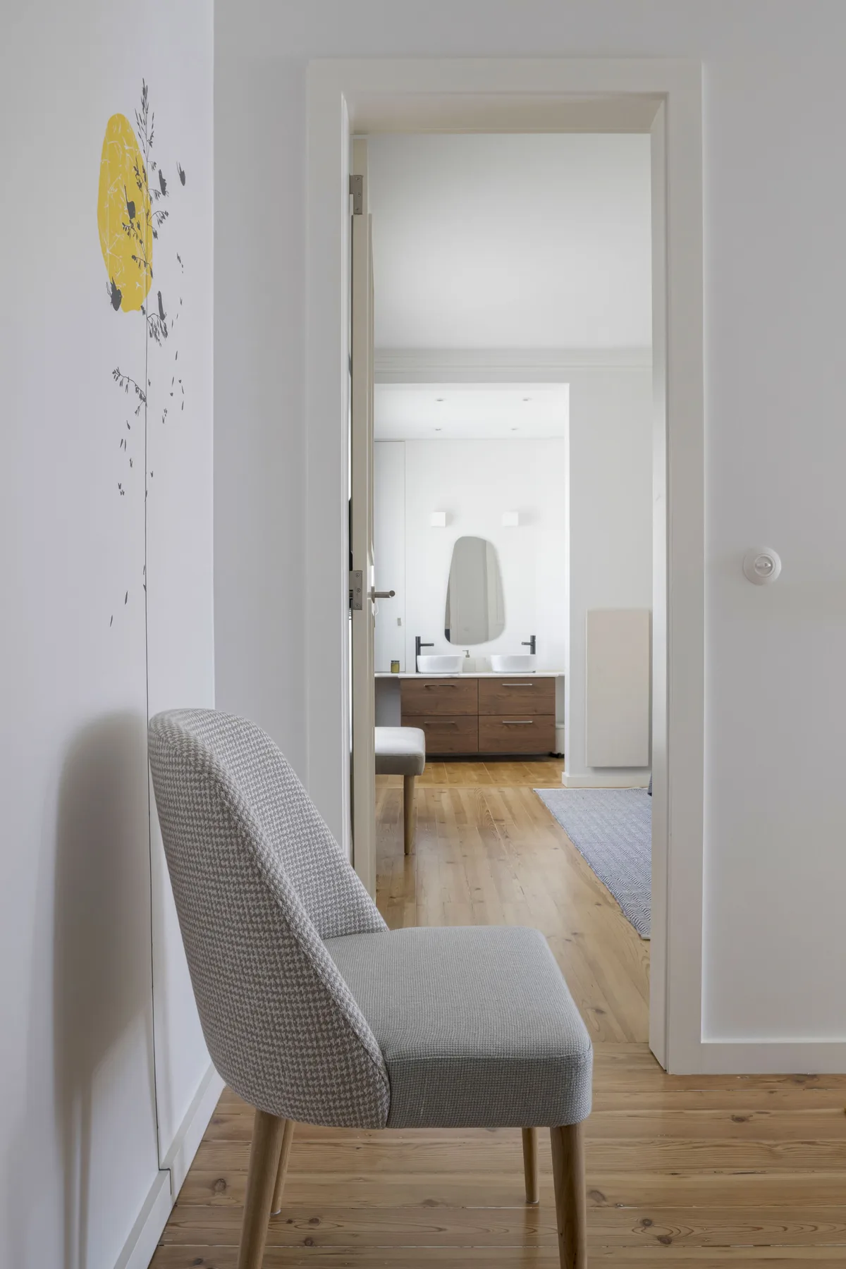Un pasillo interior luminoso da a un baño, con una silla tapizada gris en primer plano.