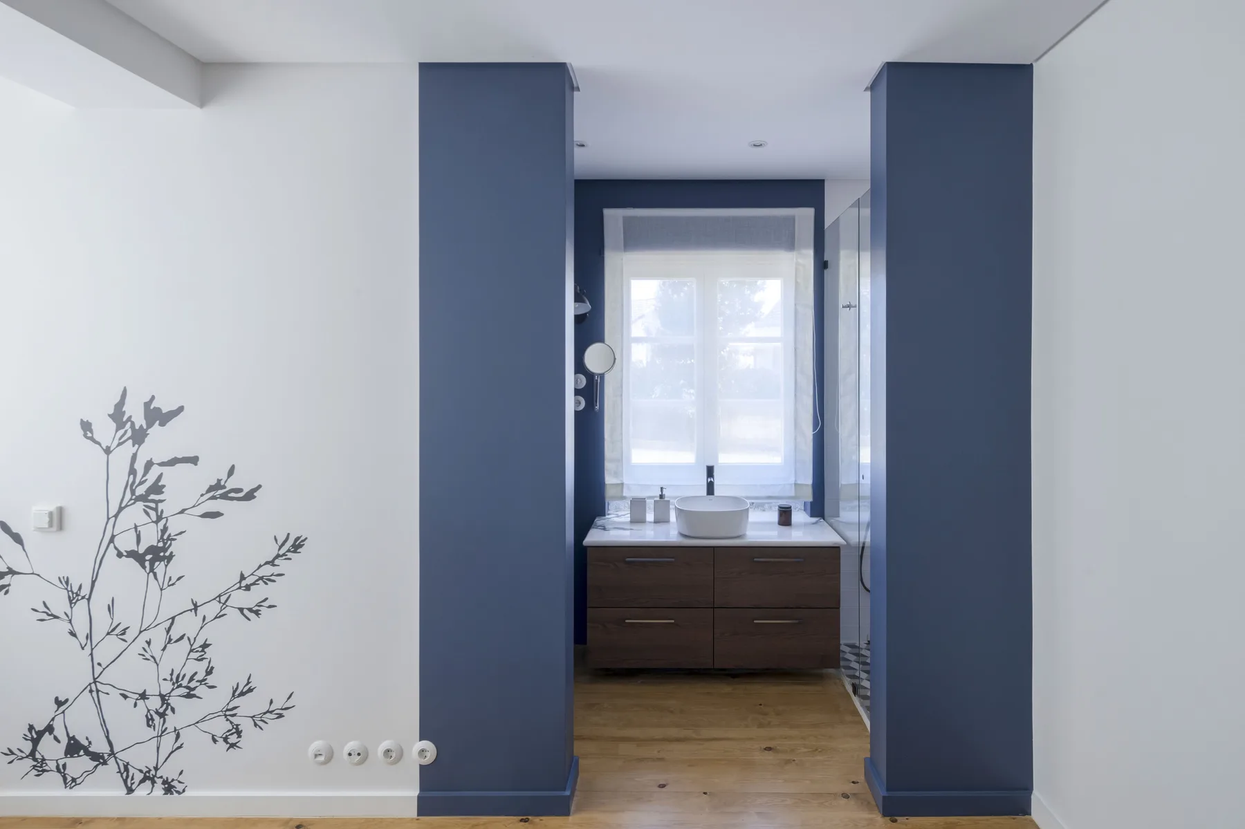 Un lavabo de baño está bajo una ventana al final de un pasillo con paredes azules.