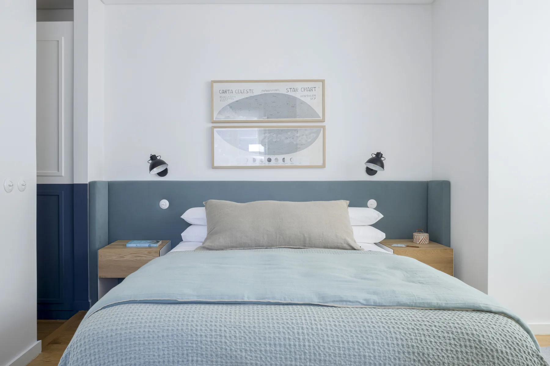 Un dormitorio luminoso con una cama tendida y láminas enmarcadas sobre el cabecero.