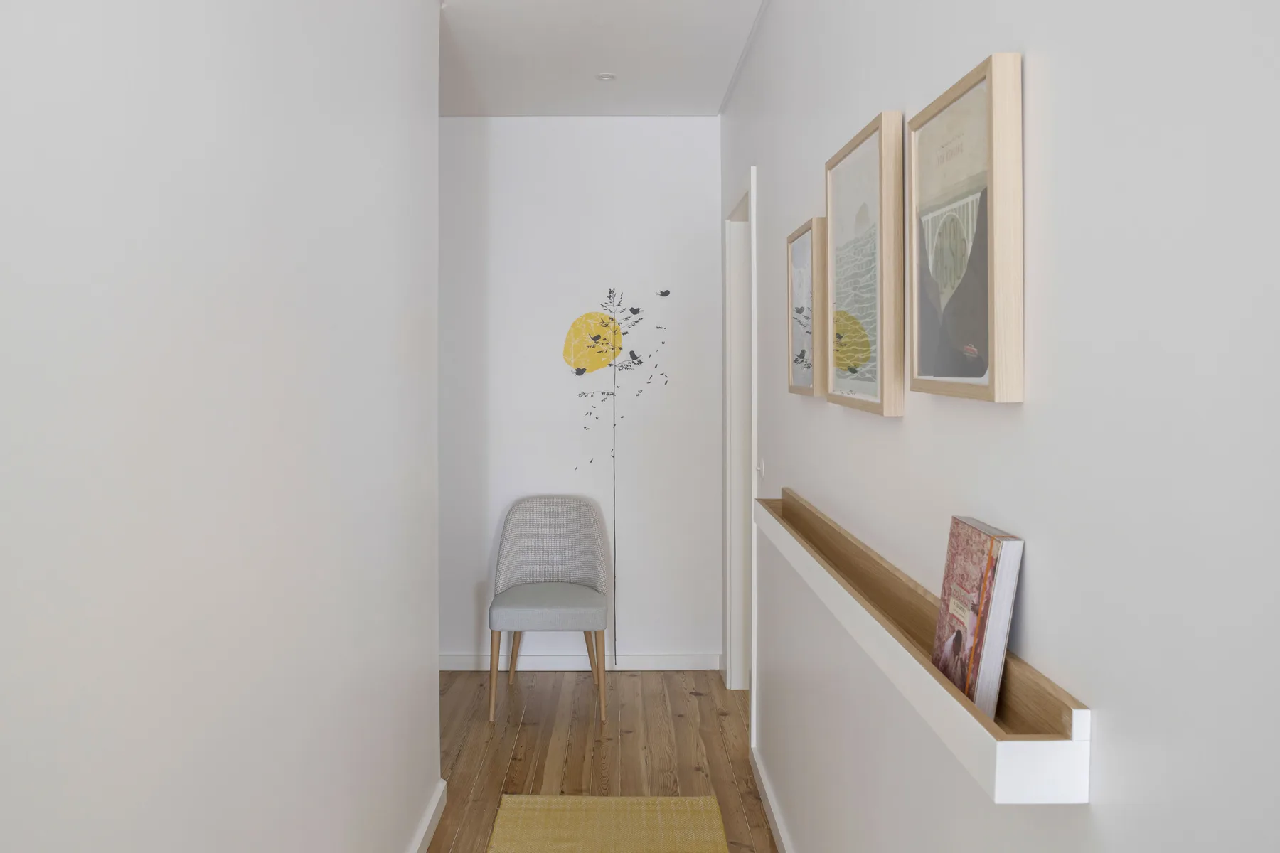 Un pasillo estrecho con cuadros, un estante de pared y una silla al fondo.