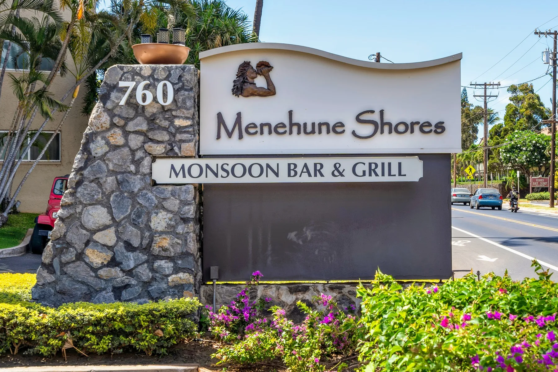 Un letrero junto a la carretera indica Menehune Shores y Monsoon Bar & Grill, con plantas floridas delante.