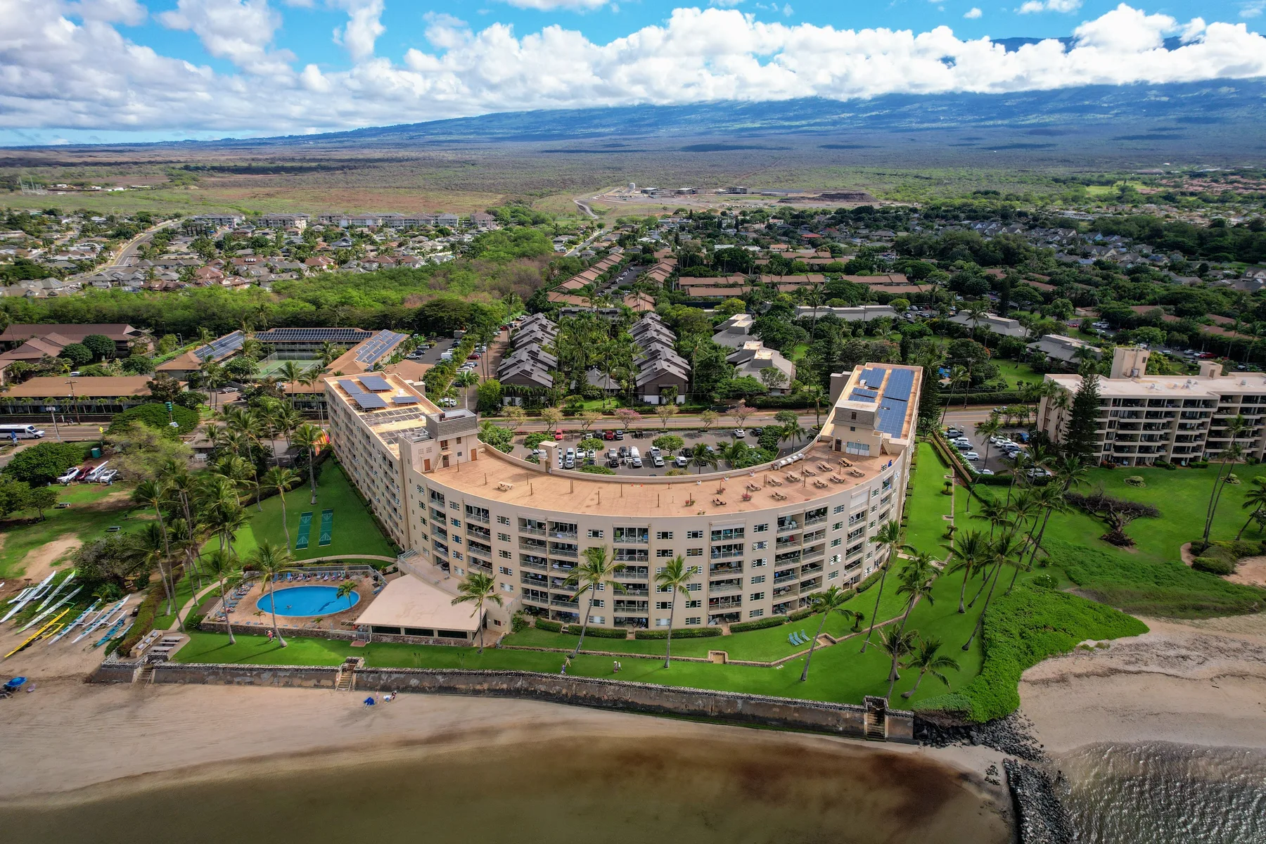Una vista aérea muestra un edificio curvo junto a la playa, con una piscina y jardines.