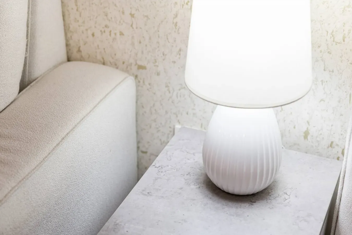 Una mesa de noche con una lámpara blanca junto a una cama tapizada.
