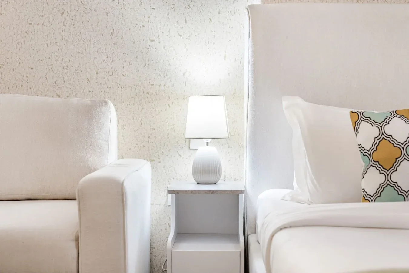 Un dormitorio con una cama blanca, un asiento tapizado y una lámpara junto a la cama.