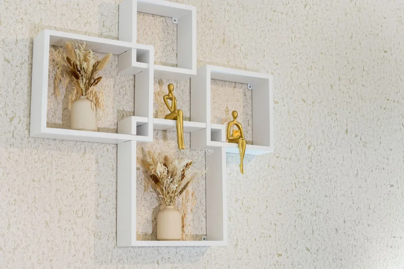Una composición de pared con estantes geométricos blancos, plantas secas y pequeñas figuras doradas.