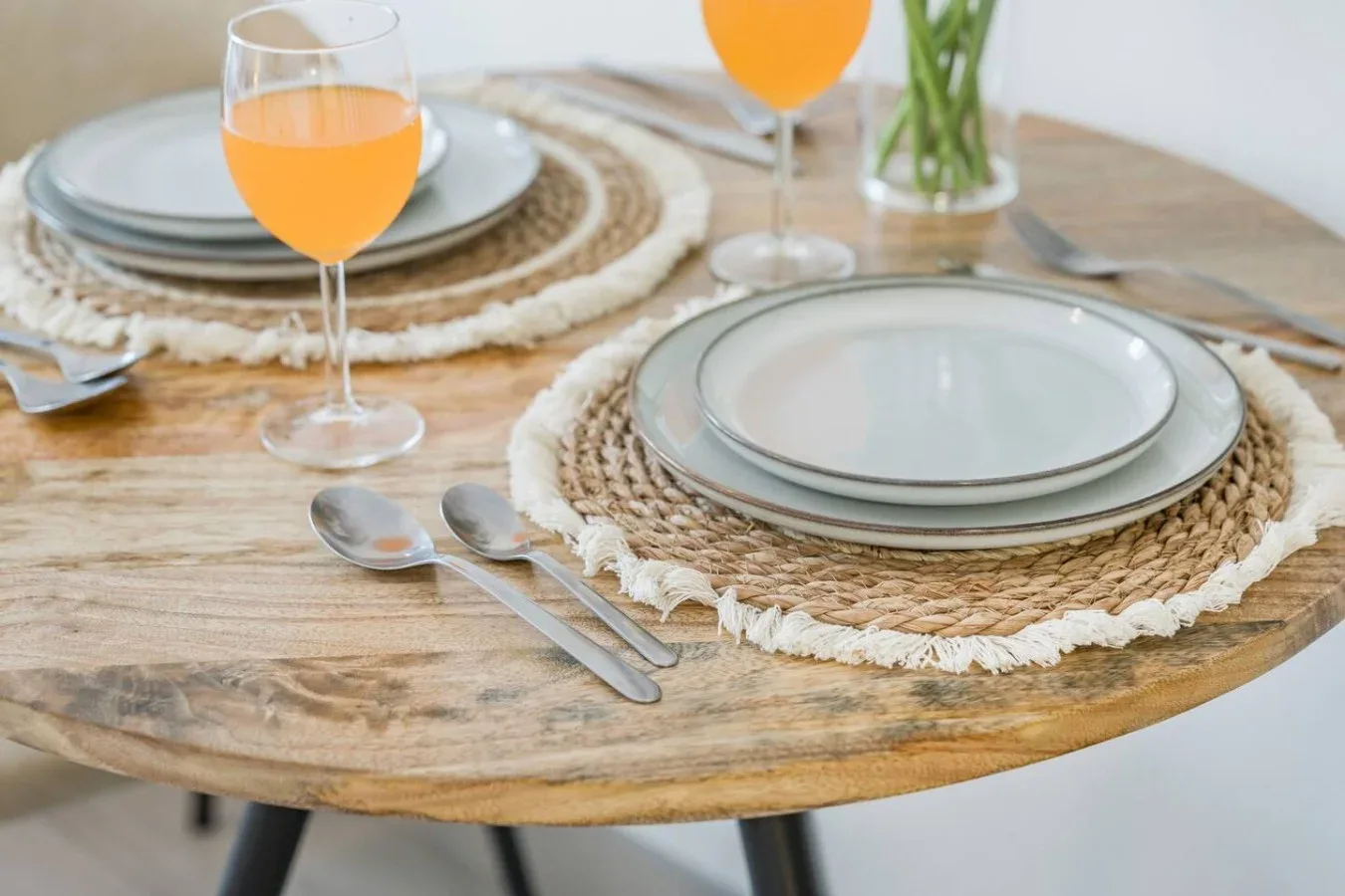 Una mesa de comedor está puesta con platos, individuales tejidos, cubiertos y bebidas anaranjadas.