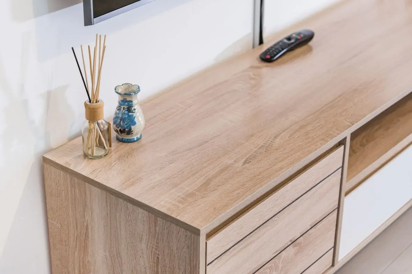 Un mueble de madera clara para televisor está bajo una pantalla montada en la pared, con pequeños adornos encima.