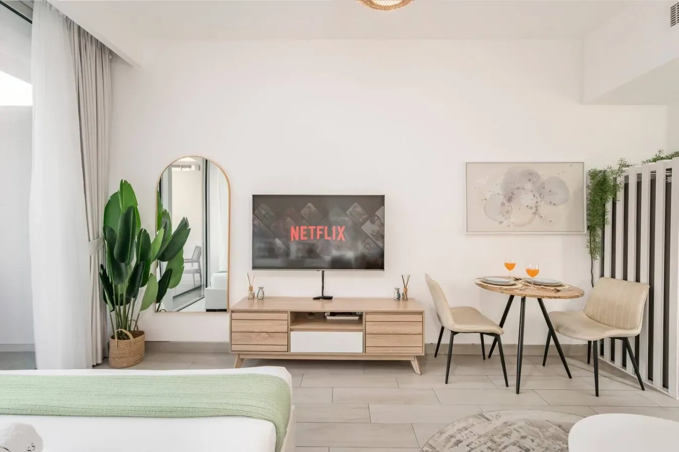 Un dormitorio luminoso con televisor, mueble bajo de madera y una pequeña mesa de comedor.