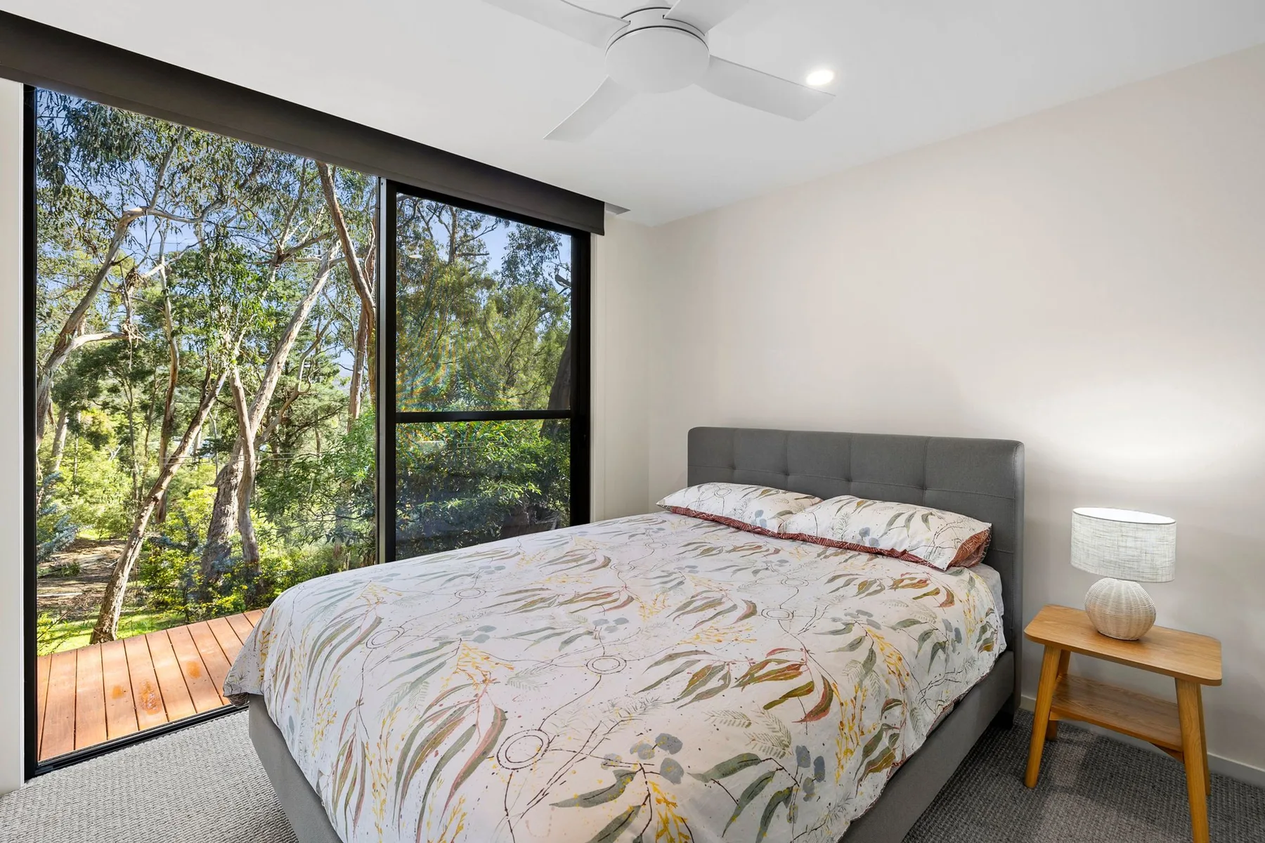 Un dormitorio con una cama junto a grandes ventanales con vistas a árboles y vegetación.