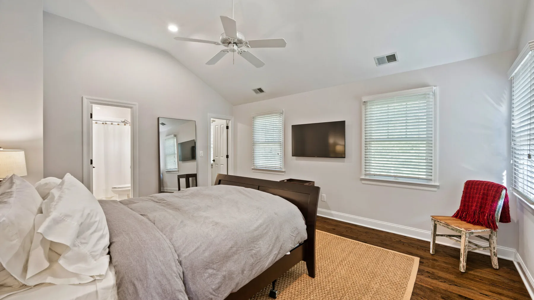 Un dormitorio luminoso con una cama, un televisor en la pared y una silla junto a la ventana.