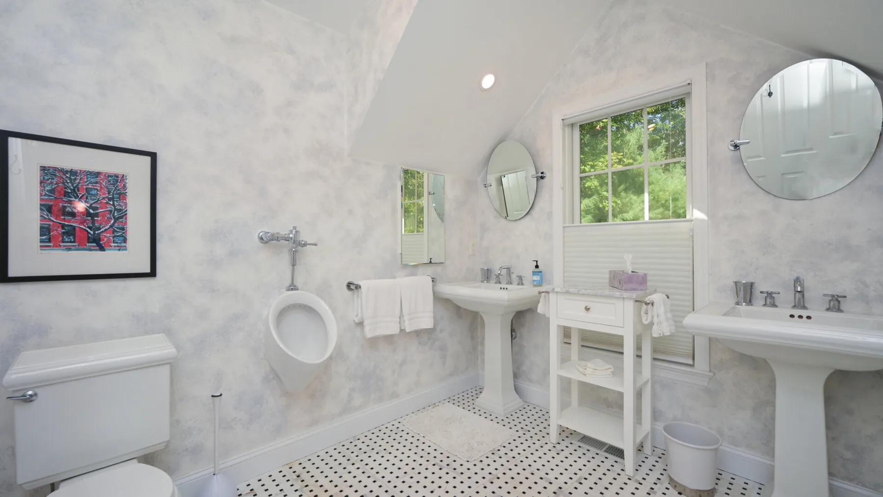 Un baño luminoso con lavabos de pedestal, un inodoro y una ventana.