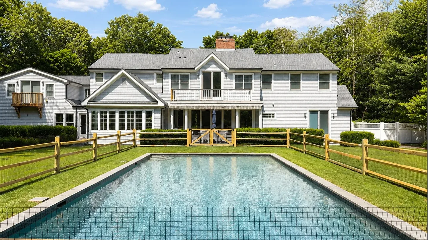 Una casa blanca grande se encuentra detrás de una piscina exterior y un césped cercado.