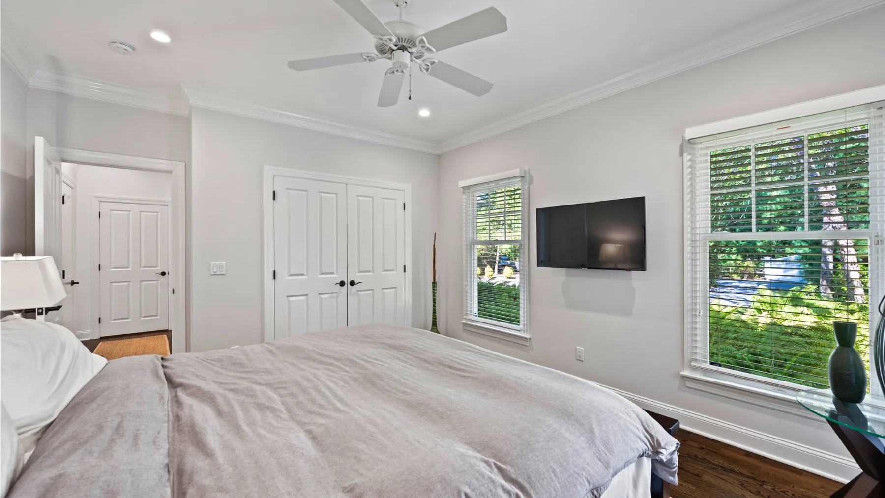 Un dormitorio con cama, ventilador de techo, televisor y ventanas con persianas.