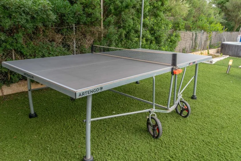 An outdoor table tennis table stands on a grassy lawn beside dense greenery.