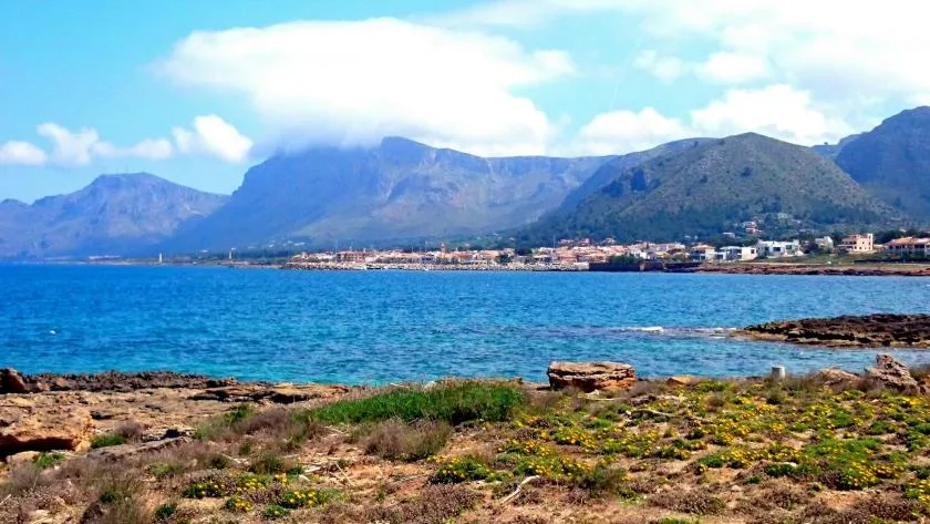 A coastal landscape with blue water, rocky shoreline, and mountains in the distance.